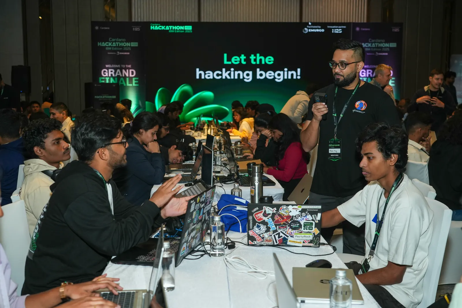 A packed hackathon floor under a 'Let the hacking begin' banner
